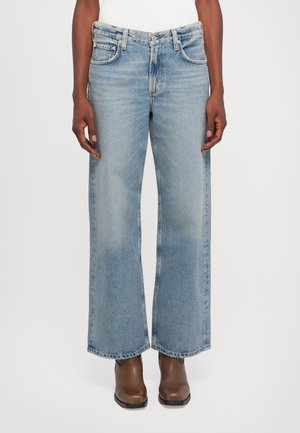 Person trägt hellblaue Jeans mit weitem Bein und braune Stiefeletten vor hellem, einfarbigem Hintergrund.