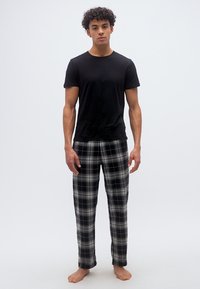 Νεαρός άνδρας που στέκεται ξυπόλυτος, φορώντας μαύρο μπλουζάκι και καρό πυτζάμες σε λευκό φόντο.