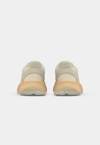 Beige atletikksko med myk mesh-overdel, buet design og en tan gummisåle. Har polstrede krager og teksturerte detaljer på hælene.