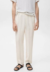 Pantalons beige clair avec des rayures verticales, arborant une coupe lâche et une texture lisse. Portés avec une chemise blanche unie et des sandales noires.