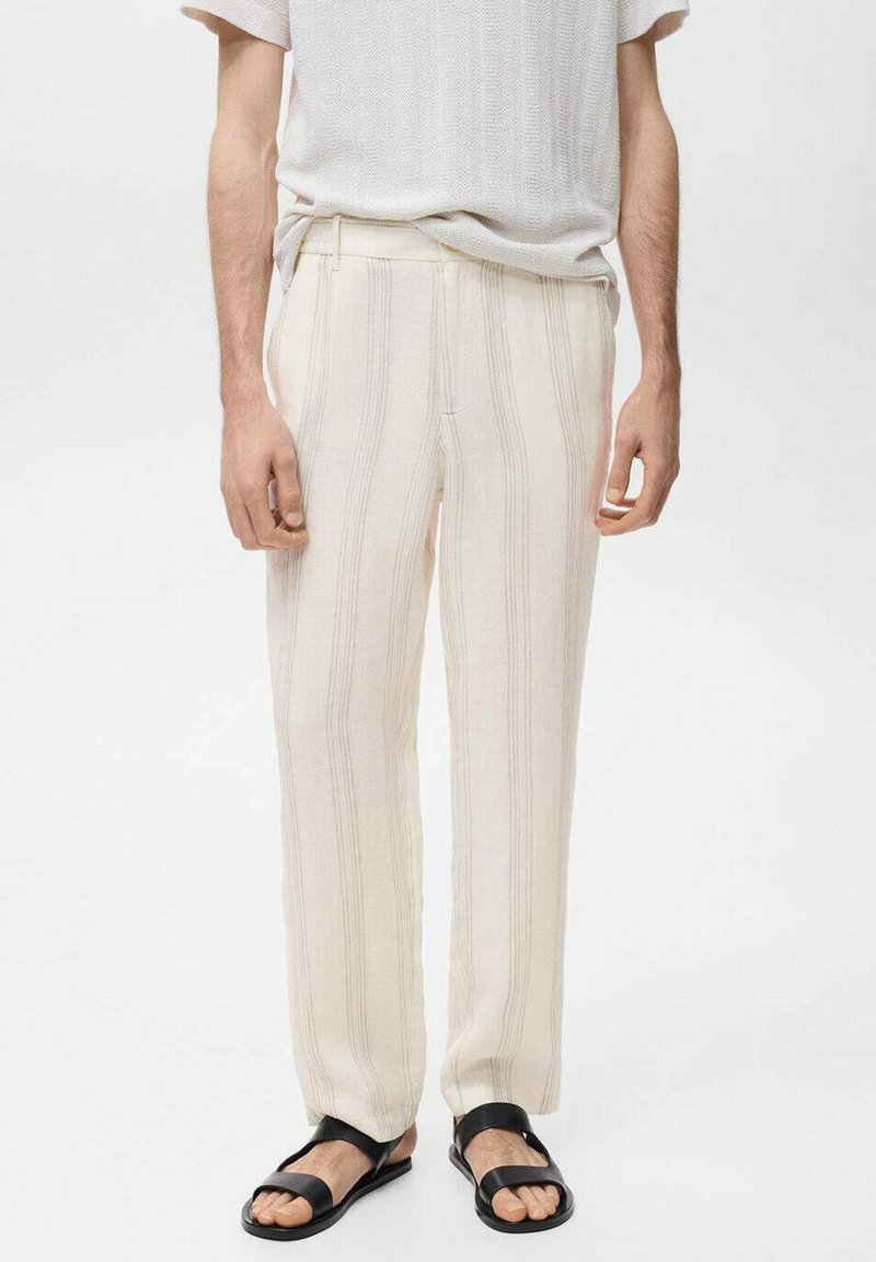Pantalons beige clair avec des rayures verticales, arborant une coupe lâche et une texture lisse. Portés avec une chemise blanche unie et des sandales noires.
