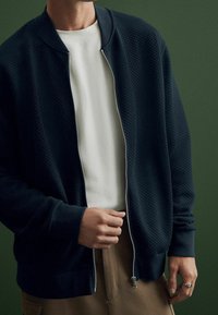 Bomber bunda s námořnickým vzorem se zipem vpředu, ribovými manžetami a límcem, nošena přes bílou košili na zeleném pozadí.