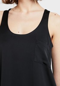 Top senza maniche nero con scollo a barca e una piccola tasca sul petto. Tessuto morbido e leggero con una vestibilità rilassata e un design minimal.