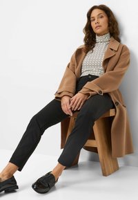 Ein brauner Wollmantel, ein gemusterter Rollkragenpullover, schwarze Slim-Fit-Hosen und schwarze Schuhe liegen auf einem Holzstuhl vor einem schlichten Hintergrund.