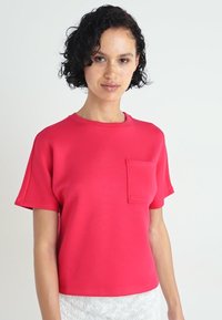 T-shirt ajusté à manches courtes de couleur rose vif, avec un col rond et une poche plaquée sur le côté gauche. Texture du tissu lisse.