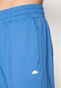 Blå sportsbukser med et struktureret stof, elastisk talje, sidelommer og en hvid logo-accent på fronten.