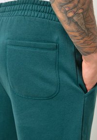 Tyrkysové tepláky mají elastický pás, boční kapsy a zadní nášivkovou kapsu s hladkou texturou a pečlivým šitím.