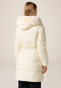 Pufferjacka i krämfärg med en huva av konstpäls. Har midjebälte, quiltad design och långa ärmar som ger värme och skydd.