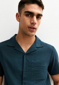 Camisa de manga corta en verde azulado oscuro con un patrón texturizado, que cuenta con un cuello con muesca, cierre de botones y un bolsillo en el pecho.