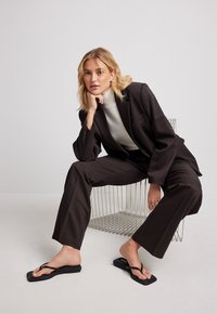 Veste de costume brun ajustée et pantalons à jambes larges, associés à un pull à col roulé gris clair. Des tongs noires complètent la tenue. Fond neutre.