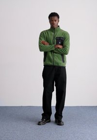 Giacca in pile verde con zip frontale, dotata di una tasca nera e dettagli scuri, abbinata a pantaloni e scarpe nere. Design semplice.