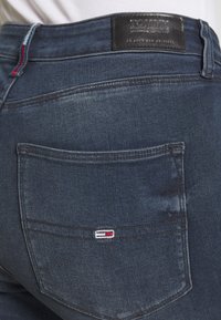 Jean de denim azul oscuro con una textura suave, que presenta un parche con el logo de Tommy Jeans en la parte trasera y acentos de costura en rojo, blanco y azul.