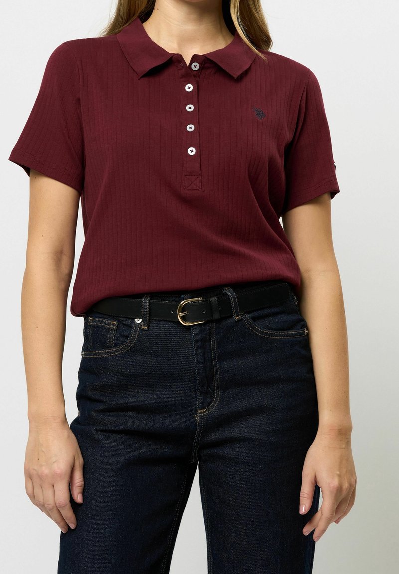 Femme portant un polo bordeaux à manches courtes avec des boutons blancs, rentré dans un jean bleu foncé et une ceinture noire avec une boucle dorée.