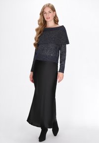 Pull en maille pailleté bleu marine avec un superposé asymétrique, associé à une jupe maxi en satin noire et des bottines noires. Texture lisse.