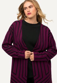 Cardigan viola con motivi geometrici neri, design aperto frontale, maniche lunghe e texture morbida. Indossato sopra un top nero.