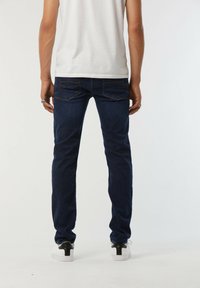 Un jean bleu foncé avec une coupe slim, présentant des coutures contrastantes, quatre poches et une texture lisse, associé à des baskets blanches.