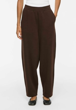 Personne portant un pantalon ample marron foncé avec une ceinture élastique et des poches, associé à des chaussures plates noires et un haut blanc.