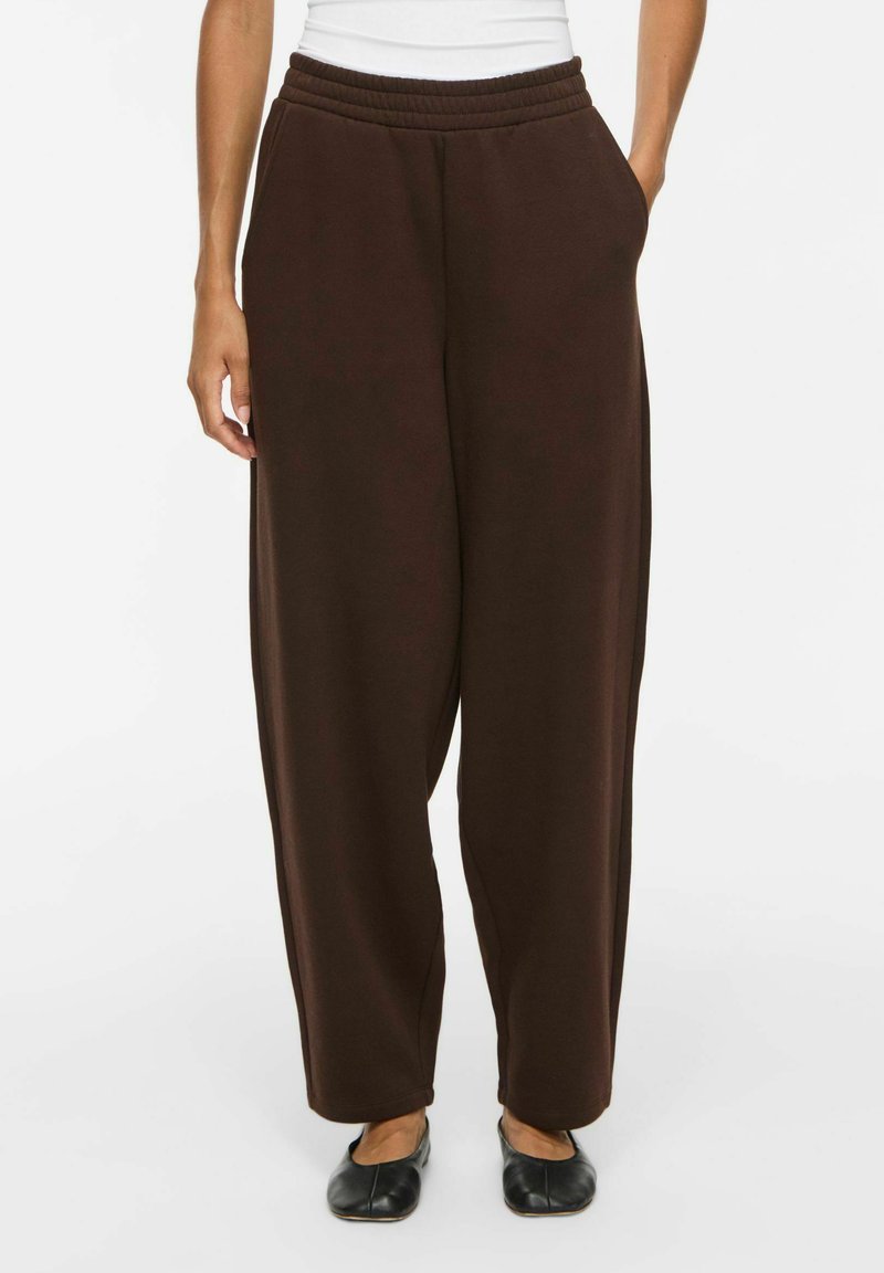 Personne portant un pantalon ample marron foncé avec une ceinture élastique et des poches, associé à des chaussures plates noires et un haut blanc.