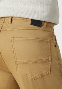 Tan cotton pants with a back pocket, featuring topstitching and a black leather brand label. Smooth texture with a structured fit.