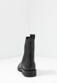 Botín negro de cuero hasta el tobillo con una lengüeta, paneles laterales elásticos y un tacón bajo, visto desde atrás sobre una superficie blanca.