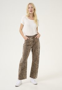 Pantalons à larges jambes en tissu beige et marron à motif léopard, associés à un t-shirt blanc simple et des baskets blanches. Coupe décontractée et style casual.