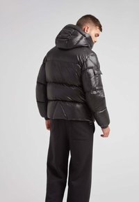 Giacca a vento nera con cappuccio, caratterizzata da una finitura lucida, un design trapuntato e una tasca sul braccio. Abbinata a pantaloni scuri.