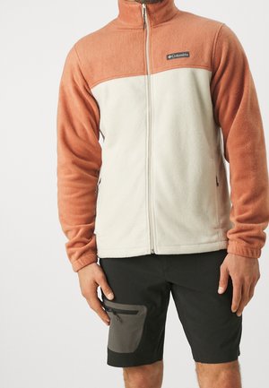 Man draagt een tweekleurige Columbia fleecejas met een volledige rits en zwarte shorts met een ritssluitingzak, staand tegen een effen achtergrond.