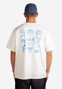 Bílá bavlněná tričko s modrou grafickou grafikou na zadní straně, zahrnující tvary jako hory, vlny a text "DILLY PARADISE RESORT."