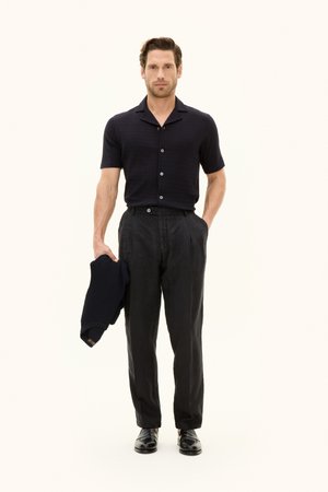 Homme portant une chemise noire à manches courtes boutonnée, un pantalon noir et des chaussures noires, tenant un pull noir plié dans une main.