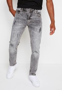 Blekta grå denimjeans med slitna detaljer, en snäv passform och fem fickor, i kombination med vita sneakers på en neutral bakgrund.