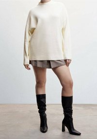 Pull en maille côtelée couleur crème associé à un short marron à motifs et à des bottes à talons hautes en cuir noir jusqu'aux genoux.