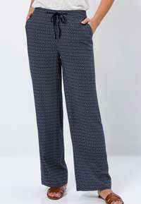 Vrouw in marineblauwe, gestreepte, wijde broek met een trekkoord in de taille en bruine instap sandalen, staande met de handen in de zakken.