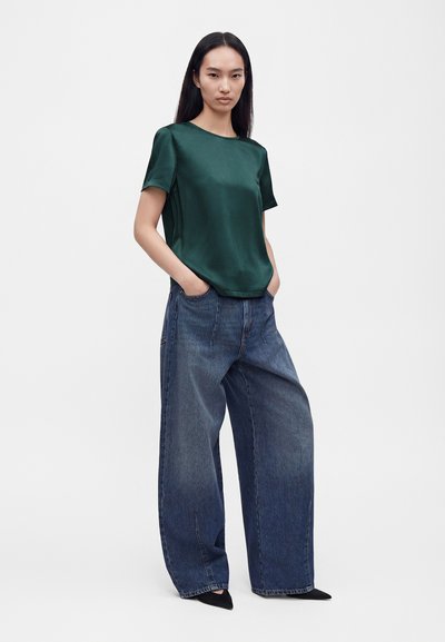 Femme aux longs cheveux noirs portant un haut en satin vert foncé à manches courtes et un jean large bleu ample, debout les mains dans les poches.