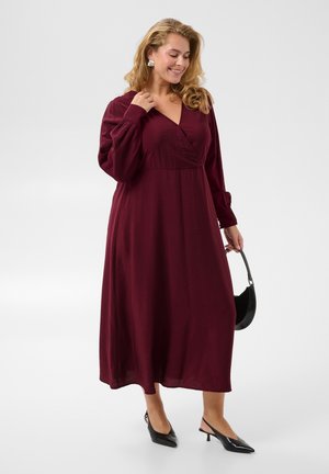 Femme en longue robe bordeaux tenant un sac à main noir, portant des talons slingback noirs et des boucles d'oreilles argentées, souriante et regardant vers le bas.