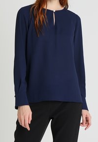 Blouse bleu marine à manches longues avec un col rond et une petite ouverture en forme de goutte, dotée de poignets boutonnés et d'un tissu lisse et léger.