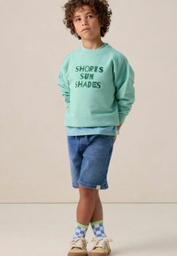 Sweatshirt vert menthe avec "SHORTS SUN SHADES" imprimé en vert foncé, associé à un short en denim et des chaussettes à carreaux colorées. Des baskets beige complètent le look.