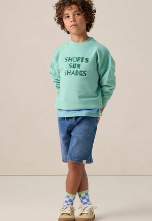 Sweatshirt vert menthe avec "SHORTS SUN SHADES" imprimé en vert foncé, associé à un short en denim et des chaussettes à carreaux colorées. Des baskets beige complètent le look.