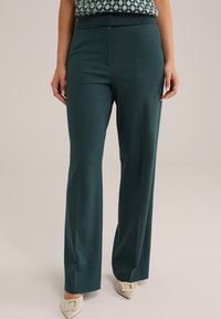 Pantalons ajustés de couleur vert foncé avec une texture lisse, une taille large et une légère évasement en bas, associés à des chaussures à bouts pointus crème.