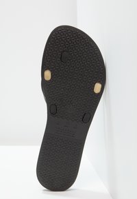 Semelle en caoutchouc noir avec un motif texturé, deux tampons de préhension circulaires et des détails de marque en relief.