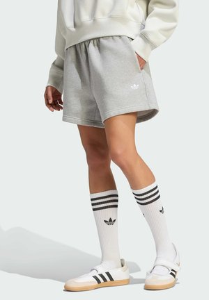 Person iført grå shorts, hvid sweatshirt, hvide Adidas-sokker med sorte striber og hvide Adidas-sneakers med sorte striber.