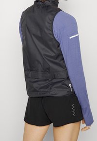 Gilet nero senza maniche con collo alto, abbinato a una maglia a maniche lunghe blu con texture. Presenta dettagli riflettenti e un design aderente.