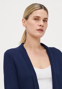 Marineblå cardigan med teksturert strikkedesign, åpen front og lange ermer, lagd over en hvit topp. Enkel og allsidig.