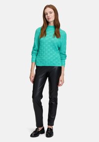 Türkiser gestrickter Pullover mit einem strukturierten Muster, kombiniert mit schwarzen Kunstlederhosen und schwarzen Loafers. Das Model steht mit Händen in den Hüften.