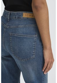 Detailní záběr osoby nosící modré džínové kalhoty s zadním kapsou a hnědou nášivkou značky "ICHI", ruka odpočívá po straně.