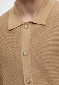 Gros plan d'un polo en maille beige avec un motif texturé et quatre boutons beiges le long de la patte de boutonnage avant.