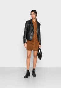 Jaqueta de motoqueiro de couro preto, vestido botão cor ferrugem com franzidos, botins pretos até o tornozelo e uma bolsa preta texturizada, contra um fundo simples.