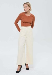 Femme aux cheveux blonds courts portant un haut long à manches rustiques, un pantalon large taille haute crème et des talons hauts noirs, se tenant les mains derrière le dos.