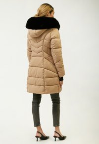 Piumino beige con un colletto in pelliccia sintetica nera, modello a zigzag, e cappuccio, abbinato a jeans scuri aderenti e scarpe con tacco nere.