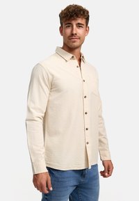 Chemise beige clair à boutons, en tissu texturé, dotée d'une poche poitrine et de boutons marron, associée à un jean bleu.
