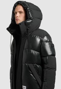 Parka noire matelassée avec une finition brillante et une capuche oversize. Dispose d'une fermeture éclair, de cordons ajustables et d'un logo sur la manche.